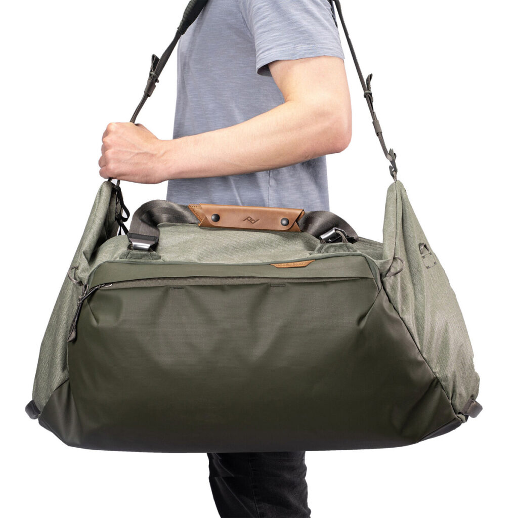Peak-Design-Travel-Duffel-65l-sage-3 Vaaleansiniseen paitaan pukeutunut henkilö kantaa Peak Design Travel Duffel 65L - Sage -laukkua, joka on suuri vihreä ja harmaa laukku, jossa on nahka-aksentteja, useita lokeroita ja musta olkahihna.