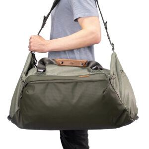 Peak-Design-Travel-Duffel-65l-sage-3 Vaaleansiniseen paitaan pukeutunut henkilö kantaa Peak Design Travel Duffel 65L - Sage -laukkua, joka on suuri vihreä ja harmaa laukku, jossa on nahka-aksentteja, useita lokeroita ja musta olkahihna.