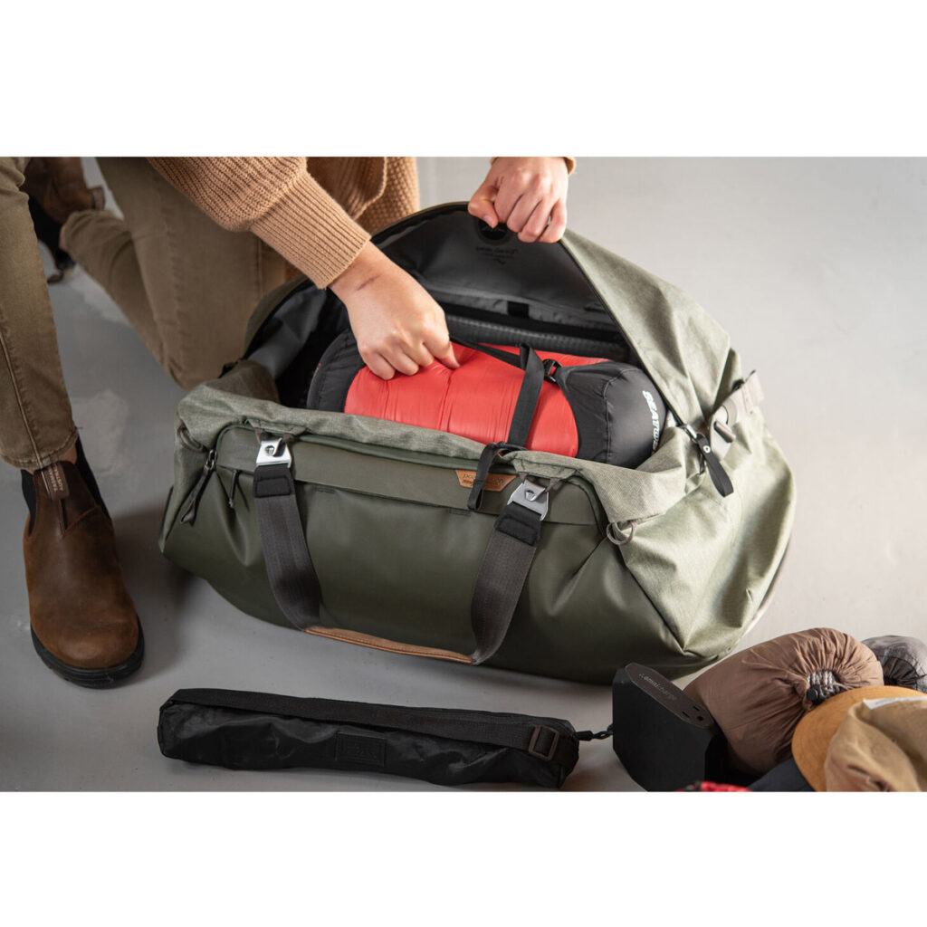 Peak-Design-Travel-Duffel-65l-sage-4 Ruskeapukuinen henkilö pakkaa punamustan makuupussin Peak Design Travel Duffel 65L - Sage -laukkuun. Erilaisia matkatarvikkeita lojuu lattialla vieressä.