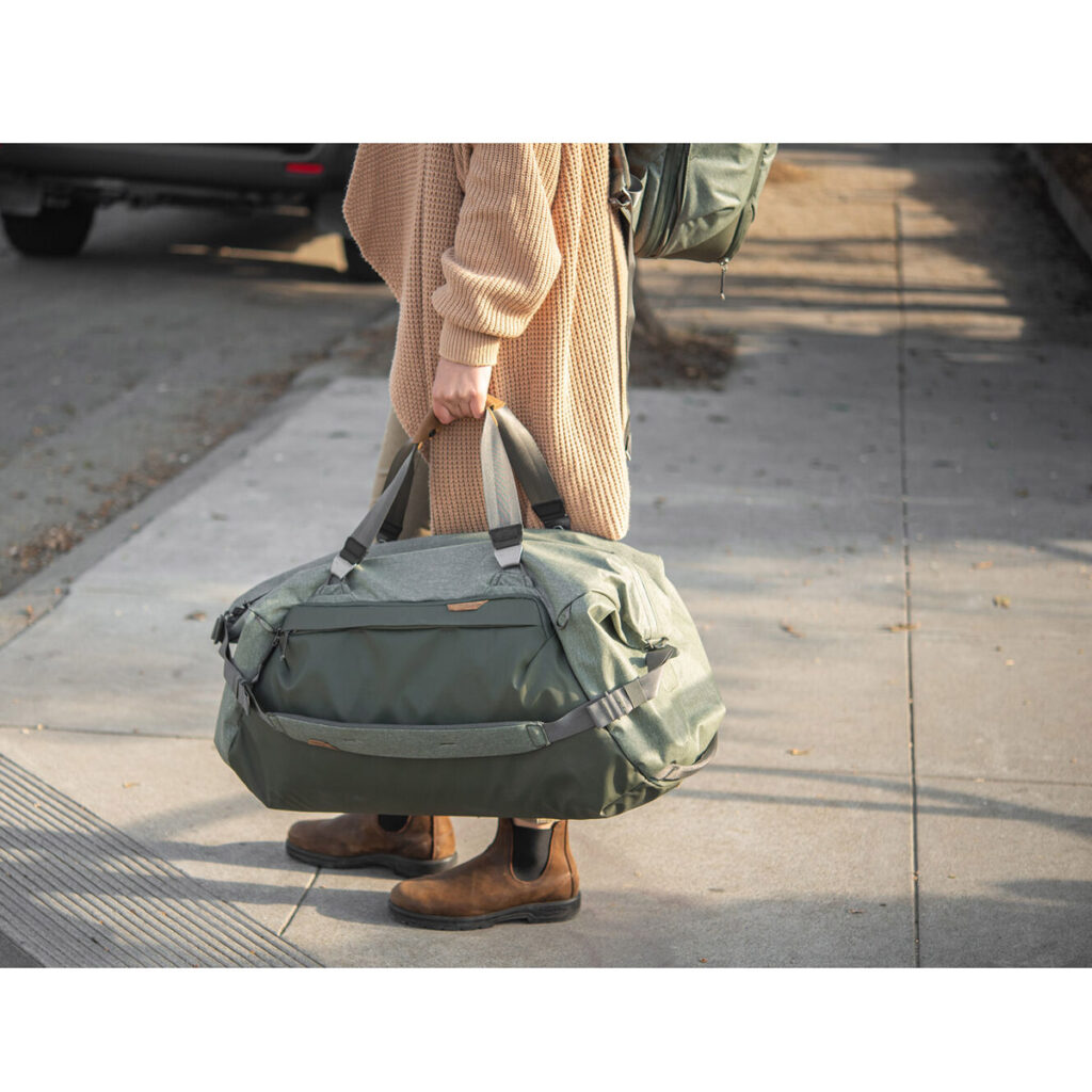 Peak-Design-Travel-Duffel-65l-sage-5 Henkilö, jolla on ruskehtava pusero, ruskeat housut ja ruskeat saappaat, seisoo jalkakäytävällä pitäen kädessään Peak Design Travel Duffel 65L - Sage -mallistoa. Hänen kasvonsa eivät näy. Taustalla on pysäköity auto ja reppu.
