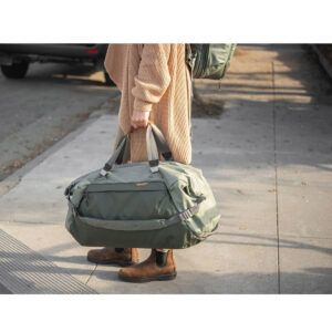 Peak-Design-Travel-Duffel-65l-sage-5 Henkilö, jolla on ruskehtava pusero, ruskeat housut ja ruskeat saappaat, seisoo jalkakäytävällä pitäen kädessään Peak Design Travel Duffel 65L - Sage -mallistoa. Hänen kasvonsa eivät näy. Taustalla on pysäköity auto ja reppu.