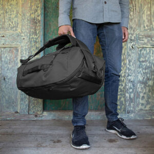 Peak-Design-Travel-Duffelpack-65l-black-08 Harmaaseen paitaan, sinisiin farkkuihin ja mustiin lenkkareihin pukeutunut henkilö seisoo puulattialla pitäen kädessään Peak Design Travel Duffelpack 65L - Black. Heidän takanaan on vanhat, sään runtelemat puuovet.