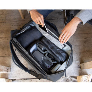 Peak-Design-Travel-Duffelpack-65l-black-10 Henkilö avaa puupinnalla olevan mustan Peak Design Travel Duffelpack 65L -matkarepun, josta paljastuu siististi pakattuja vaatteita ja järjestettyä elektroniikkaa, kuten lennokki, laturi ja kaapelit.
