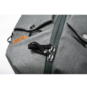 Peak-Design-Travel-Duffelpack-65l-sage-6 Lähikuva Peak Design Travel Duffelpack 65L in Sage -laukusta, harmaasta kankaasta valmistettu laukku, jossa on mustat vetoketjun vetimet, vihreä vetoketjuvuori, hopeinen laitteisto, napitettavat taskut ja osittain näkyvä rusketusruskea etiketti taskun lähellä.