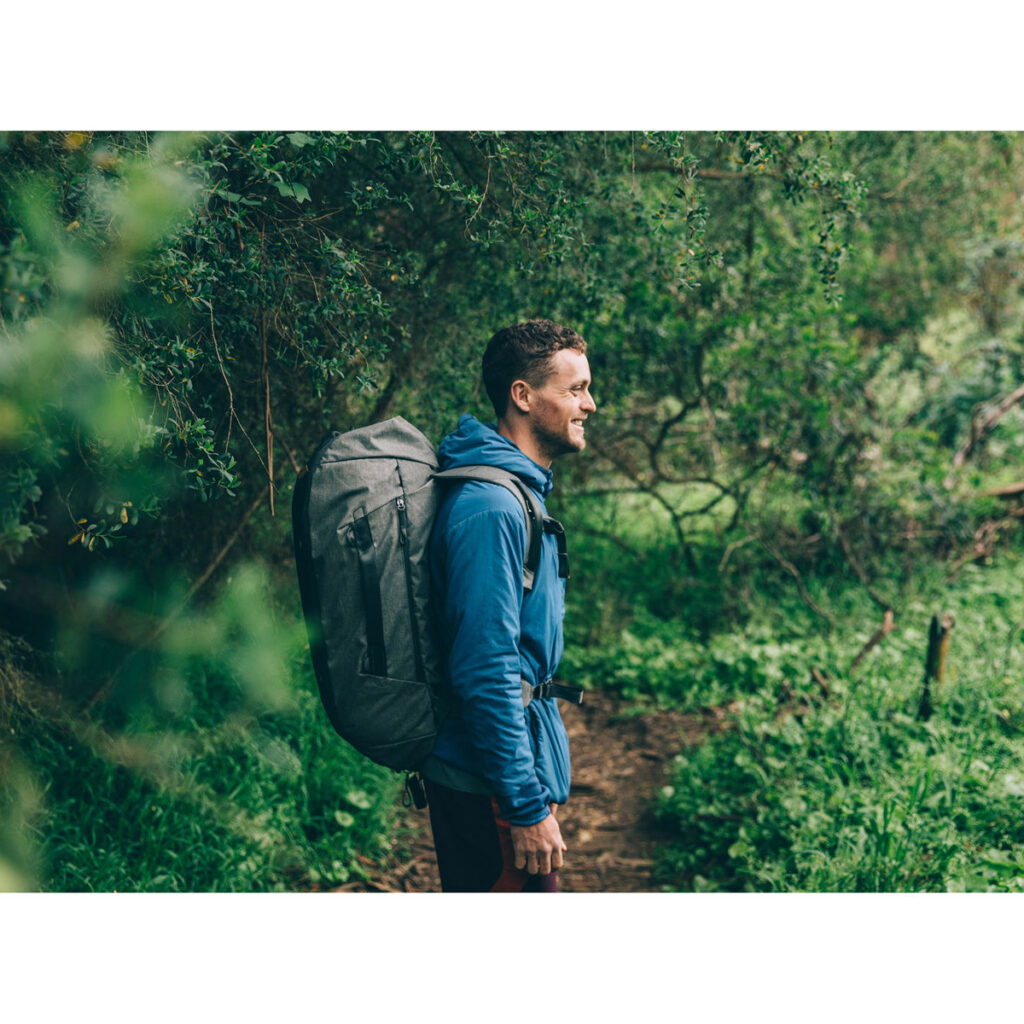 Peak-Design-Travel-Duffelpack-65l-sage-7 Siniseen takkiin pukeutunut mies, jolla on mukanaan Peak Design Travel Duffelpack 65L Sage -reppu, hymyilee seisoessaan metsäpolulla vehreiden lehtien ympäröimänä.