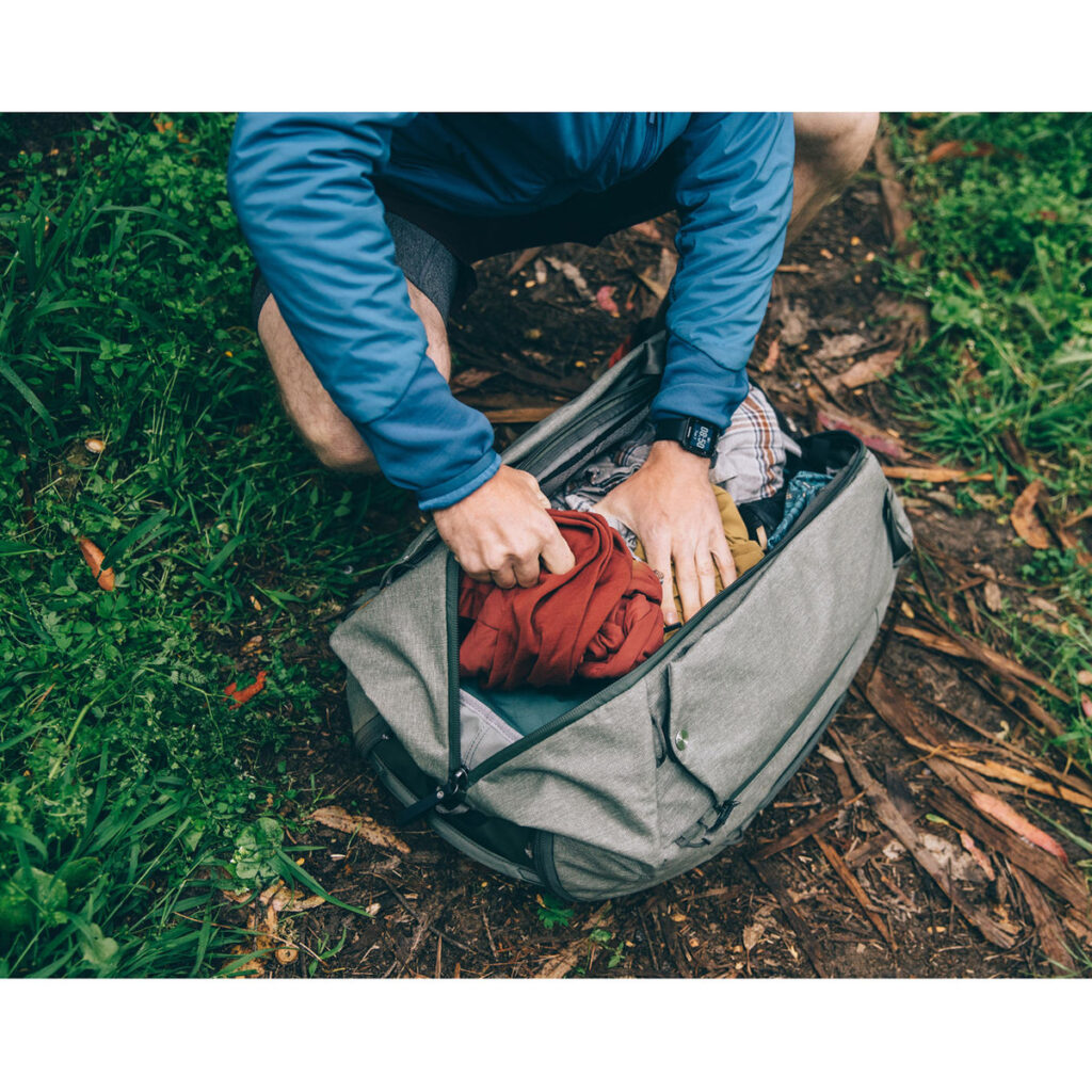Peak-Design-Travel-Duffelpack-65l-sage-8 Ulkona ruohon ja pudonneiden lehtien päällä siniseen takkiin pukeutunut henkilö polvistuu ja pakkaa vaatteita Peak Design Travel Duffelpack 65L - Sage -reppuun. Hänellä on myös kello.