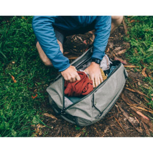 Peak-Design-Travel-Duffelpack-65l-sage-8 Ulkona ruohon ja pudonneiden lehtien päällä siniseen takkiin pukeutunut henkilö polvistuu ja pakkaa vaatteita Peak Design Travel Duffelpack 65L - Sage -reppuun. Hänellä on myös kello.