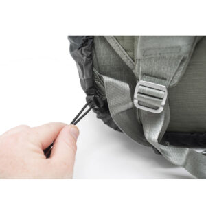 Peak-Design-Travel-Rainy-4 Lähikuva kädestä, joka vetää harmaan Peak Design Rain Fly - sadesuojan joustavaa kiristysnauhaa, reppuhihnat ja metallinen solki näkyvissä.