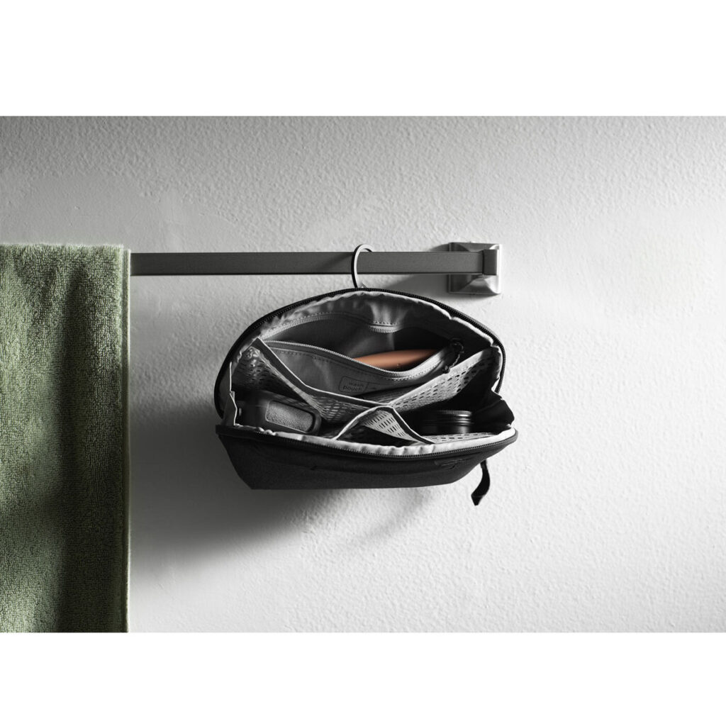 Peak-Design-Wash-Pouch-Small-Black5 Peak Design Wash Pouch Small -pesupussi, musta, roikkuu auki metallisessa pyyhetelineessä vihreän pyyhkeen vieressä, ja sen sisältä paljastuu järjestäytyneitä lokeroita, joissa on hoitotarvikkeita valkoista kuvioitua seinää vasten.