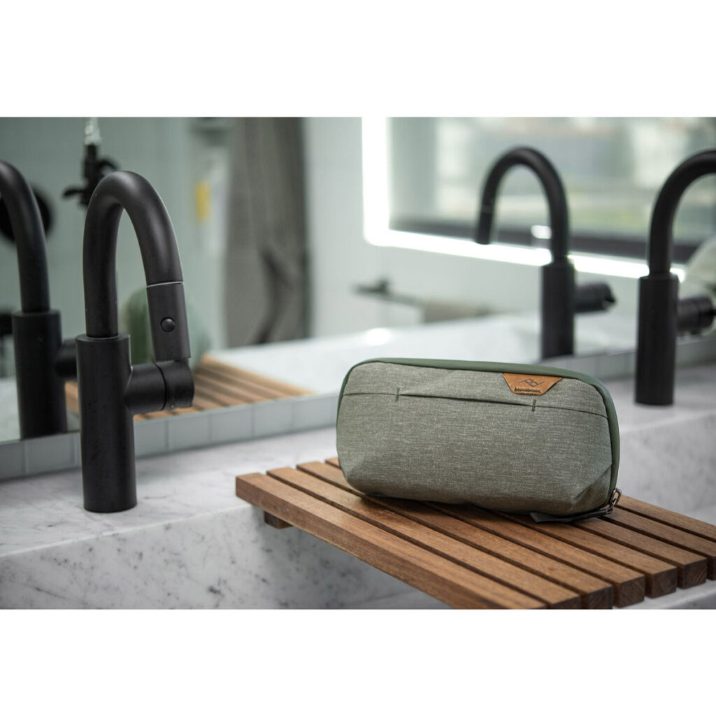 Peak-Design-Wash-Pouch-Small-Sage5 Salvia Peak Design Wash Pouch Small istuu puisella ritilätarjottimella marmorisen kylpyhuoneen pesualtaan yläpuolella, ja taustalla on musta hana ja peili.
