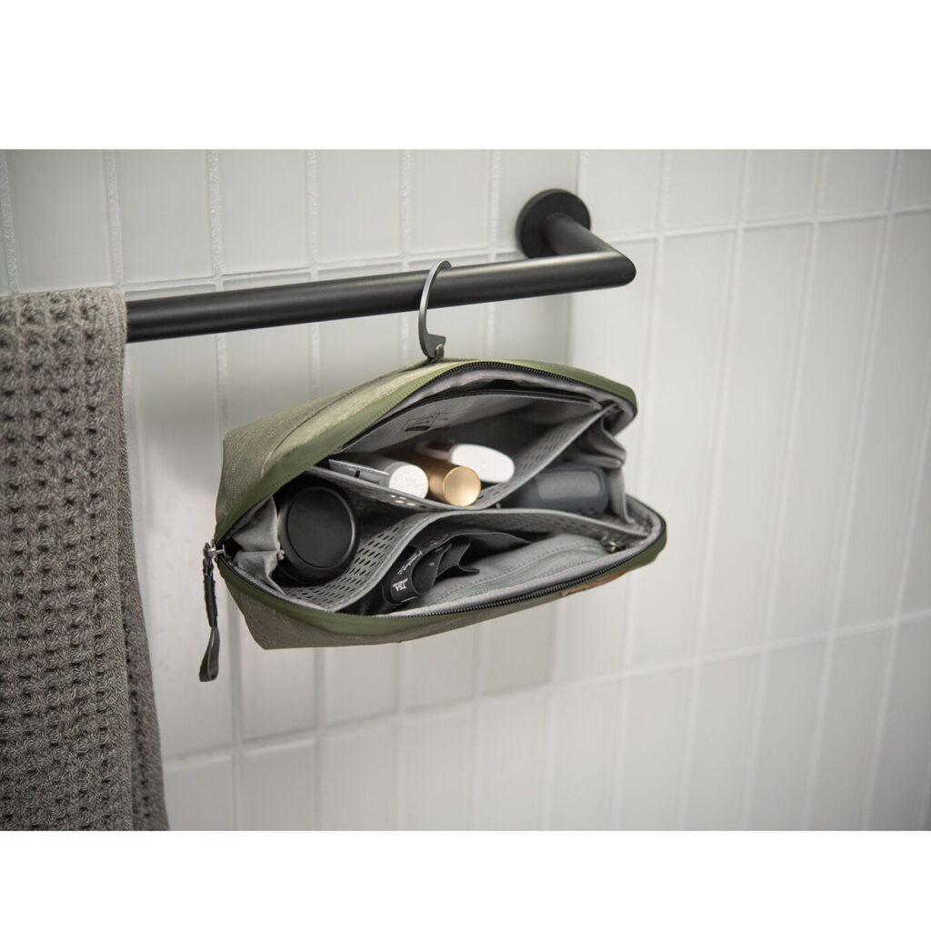 Peak-Design-Wash-Pouch-Small-Sage6 Peak Design Wash Pouch Small in Sage roikkuu auki mustasta pyyhetangosta harmaan pyyhkeen vieressä, ja sen sisällä on järjestettyjä hygieniatuotteita valkoista kaakeliseinää vasten.