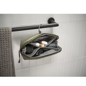 Peak-Design-Wash-Pouch-Small-Sage6 Peak Design Wash Pouch Small in Sage roikkuu auki mustasta pyyhetangosta harmaan pyyhkeen vieressä, ja sen sisällä on järjestettyjä hygieniatuotteita valkoista kaakeliseinää vasten.