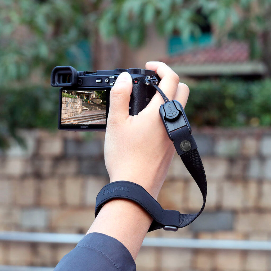 SmallRig-2398-4 Henkilö käyttää SmallRig 2398 Wrist Strap for Camera -rannehihnaa pitämään digitaalikameraa ja ottaa kuvan kivimuurista ja kameran näytöllä näkyvästä vehreästä maisemasta, jonka tausta on sumea.