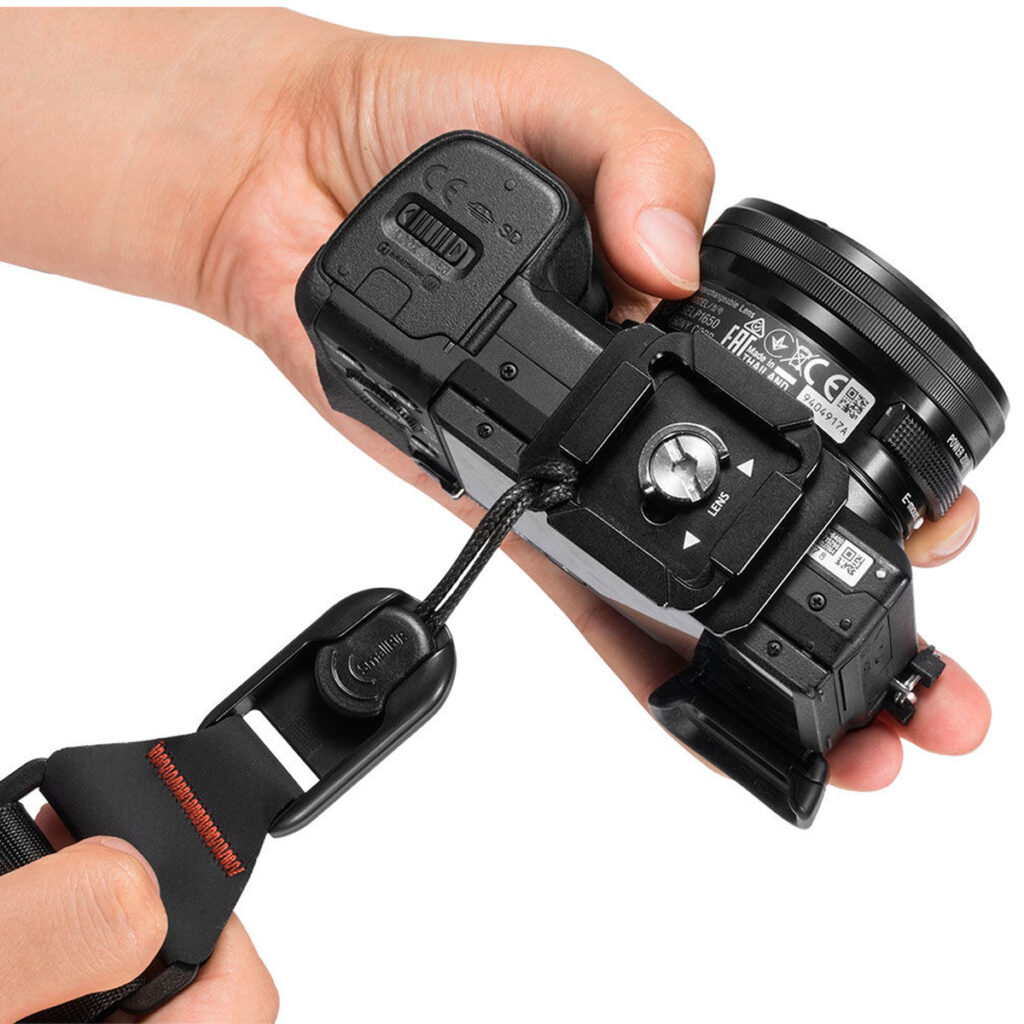 SmallRig-2428-5 Henkilö kiinnittää mustan digitaalikameran SmallRig 2428 Camera Shoulder Strap -kamerahihnalla kiinnityslevyn ja ruuvin avulla pitäen kameraa turvallisesti molemmissa käsissä.
