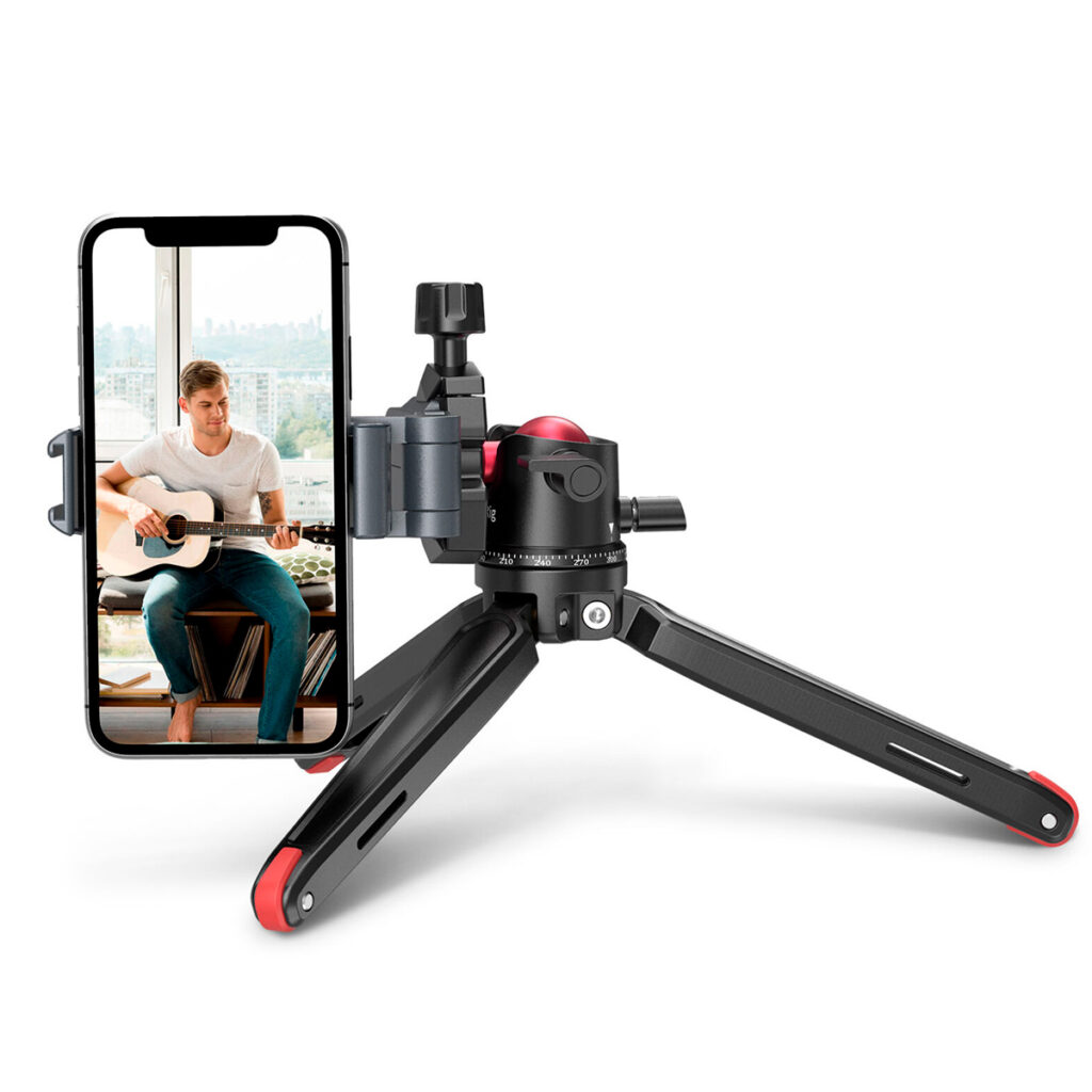 SmallRig-2664-2 A man plays acoustic guitar with a smartphone attached to a SmallRig 2664 Tabletop Minitripod with Panoramic Ballhead, and a window behind him shows a city view.