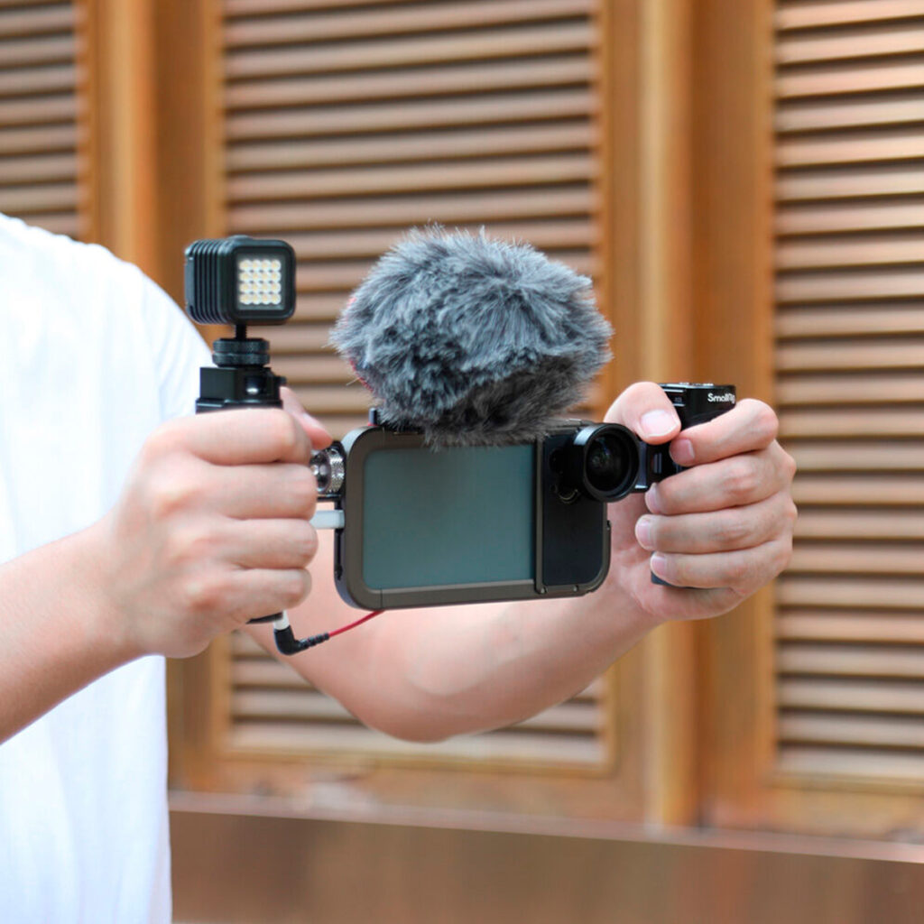 SmallRig-2772-5 Someone is holding a smartphone rig with the SmallRig 2772 Lightweight Side Handle for Smartphone Cage, equipped with a grey windscreen microphone, LED light and wide-angle lens for professional video shooting.