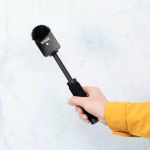 SmallRig-3182-4 Käsi keltaisessa hihassa pitää SmallRig 3182 Rode Wireless Go Handheld -haastattelulaitetta, jossa on musta vaahtomuovinen tuulisuoja ja lyhyt kahva, vaalean marmoritaustalla.