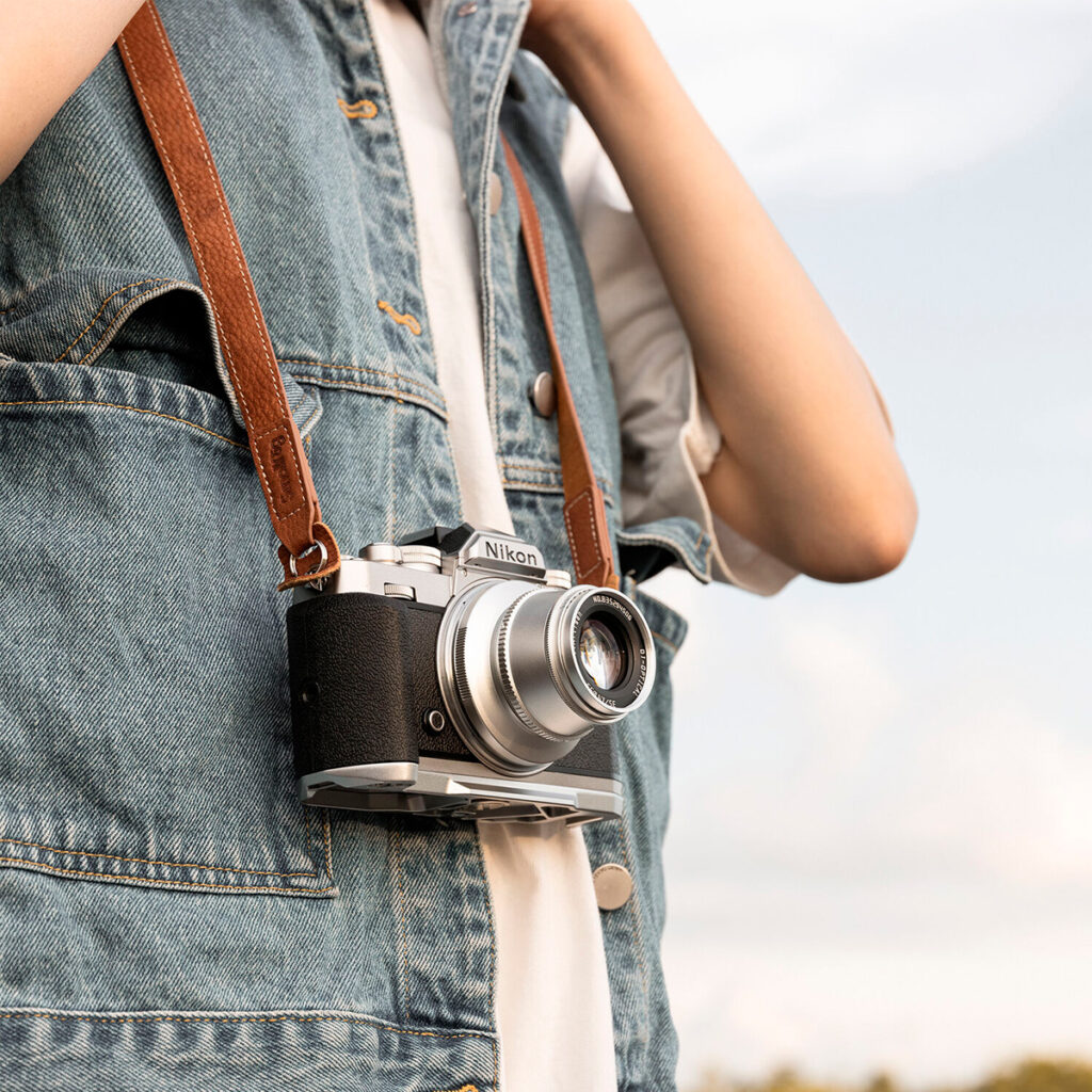 SmallRig-3485-3 A person wearing a denim vest is wearing a vintage Nikon camera around their neck with a SmallRig 3485 Leather Camera Neck Strap around their neck against a blurry sky.