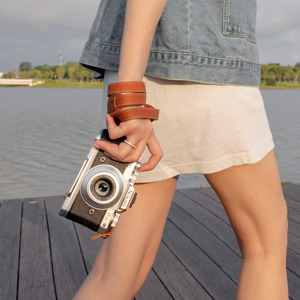 SmallRig-3485-4 A man in a white skirt and denim jacket walks along a wooden pier by a lake, holding a camera with a SmallRig 3485 Leather Camera Neck Strap. Trees and blue sky can be seen in the background.