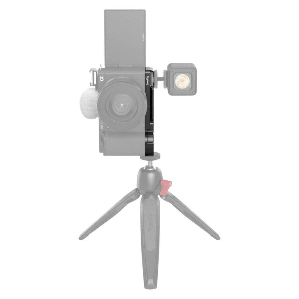 SmallRig-3523-4 Edestä katsottuna SmallRig 3523 Extension Grip for Sony ZV-E10, musta, kiinnitettynä pienellä jalustalla, ulkoinen valo kameran sivulla ja pystysuoraan asetettu kääntyvä näyttö; kamera ja jalusta näkyvät puoliksi läpinäkyvinä.