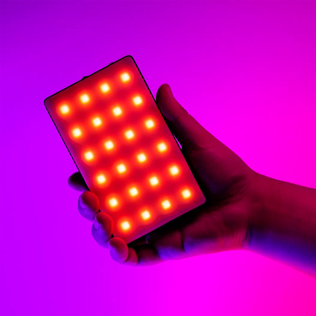 A hand holds the SmallRig 3808 RGB Videolight RM120, which shines with bright red and yellow LEDs against a vibrant pink and purple background.