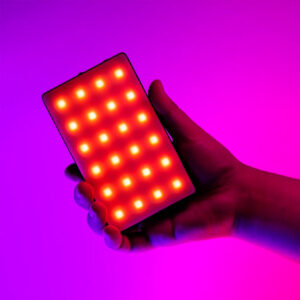 A hand holds the SmallRig 3808 RGB Videolight RM120, which shines with bright red and yellow LEDs against a vibrant pink and purple background.