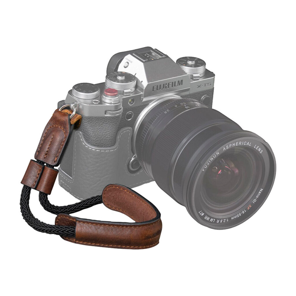 SmallRig-3926-2 SmallRig 3926 Universal Wrist Strap Vintage -rannehihna, ruskeaa nahkaa ja mustaa punosta, on esillä osittain haalistuneen Fujifilm X-T5 -kameran edessä, jossa on suuri objektiivi, joka korostaa tyylikästä rannehihnaa.