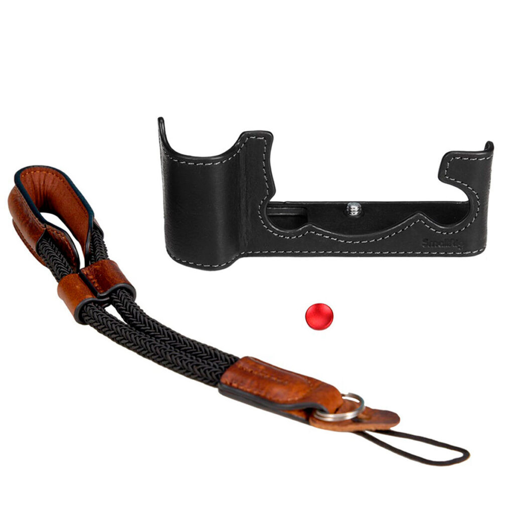 SmallRig-3927-2 SmallRig 3927 Half Case & Wrist Strap Fujifilm X-T5:lle sisältää mustan nahkaisen puolikotelon, jossa on valkoiset tikkaukset, punotun mustan ja ruskean nahkarannehihnan ja pienen punaisen painikkeen, jotka kaikki näkyvät valkoisella pohjalla.