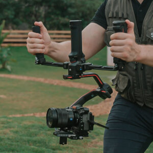 SmallRig-3954-5 Henkilö pitää SmallRig 3954 Dual Handgrip with Wireless Control for DJI RS Series -kameraa, johon on asennettu kamera ulkona ruohikolla, yllään tummat liivit ja mustat housut.