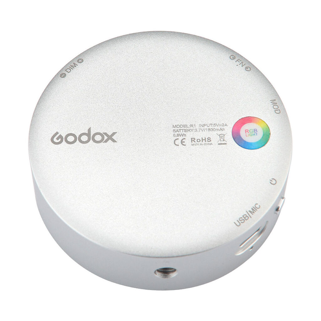 Godox_R1_003 Godox R1 on pyöreä, hopeinen laite, jonka pinnalla on merkittyjä painikkeita, portteja, RGB-valon merkkivalo ja painettu teksti sertifiointimerkinnöillä.