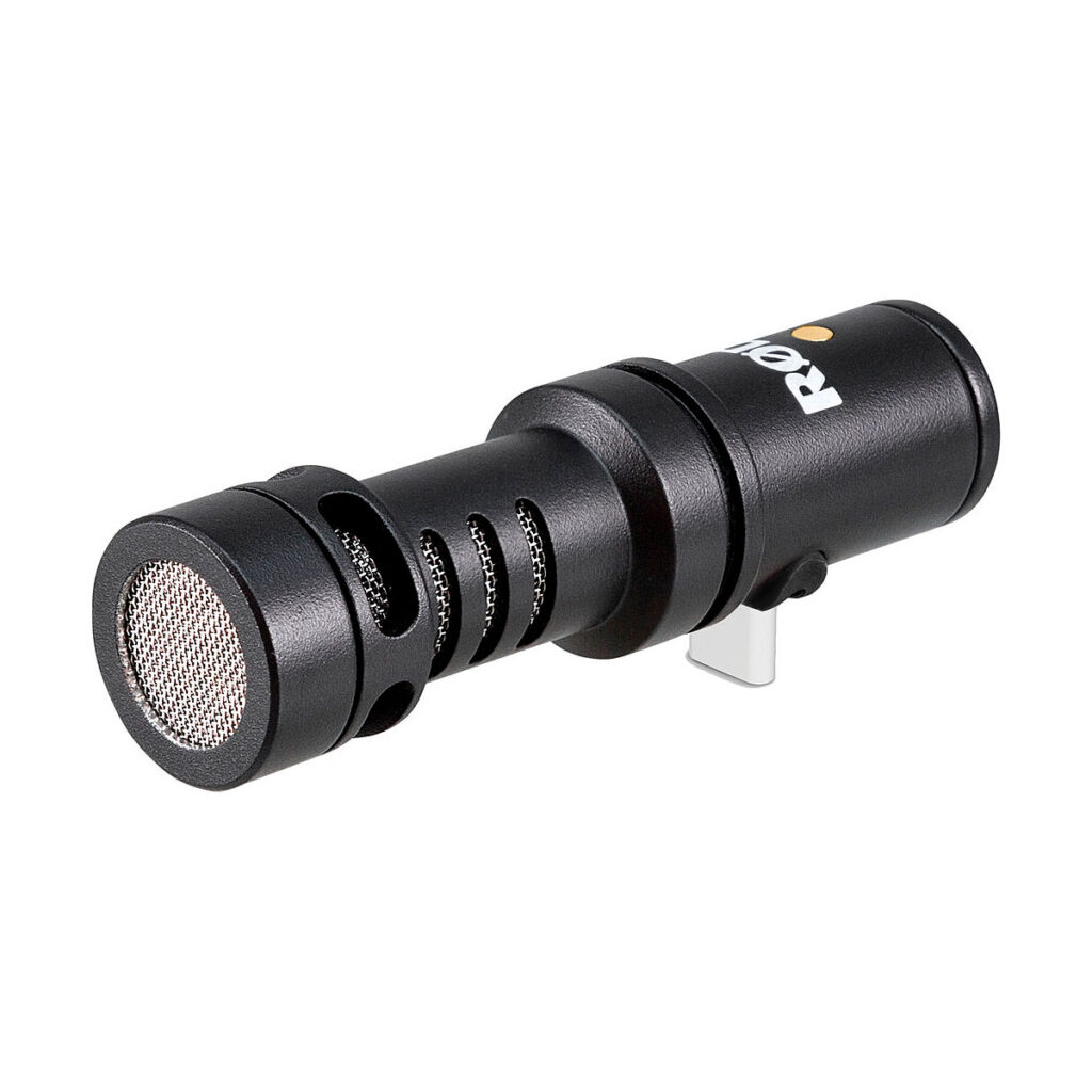 Rode_Video_Mic_Me-C_003 Musta sylinterimäinen Rode Videomic ME-C -mikrofoni, jossa on metalliverkkosäleikkö ja tuuletetut sivut, kiinnitettynä hopeiseen pidikkeeseen valkoista taustaa vasten.