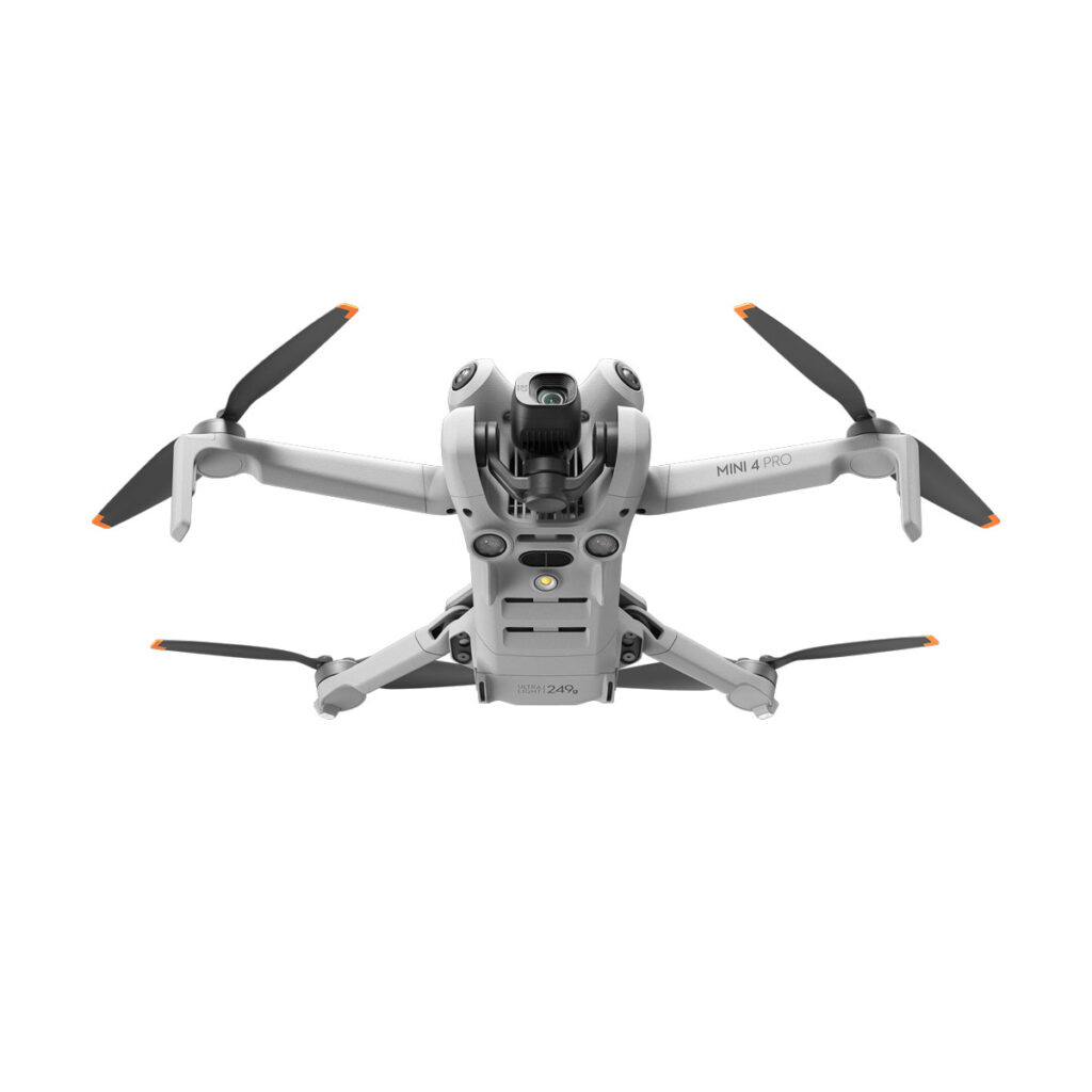 DJI_mini4pro_004 Hopeanvärinen DJI Mini 4 Pro -lennokki, jossa on neljä oranssilla kärjellä varustettua potkuria ja etukamera, näkyy lennossa valkoista taustaa vasten.