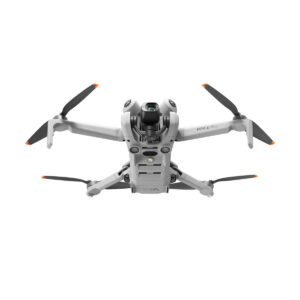 DJI_mini4pro_004 Hopeanvärinen DJI Mini 4 Pro -lennokki, jossa on neljä oranssilla kärjellä varustettua potkuria ja etukamera, näkyy lennossa valkoista taustaa vasten.