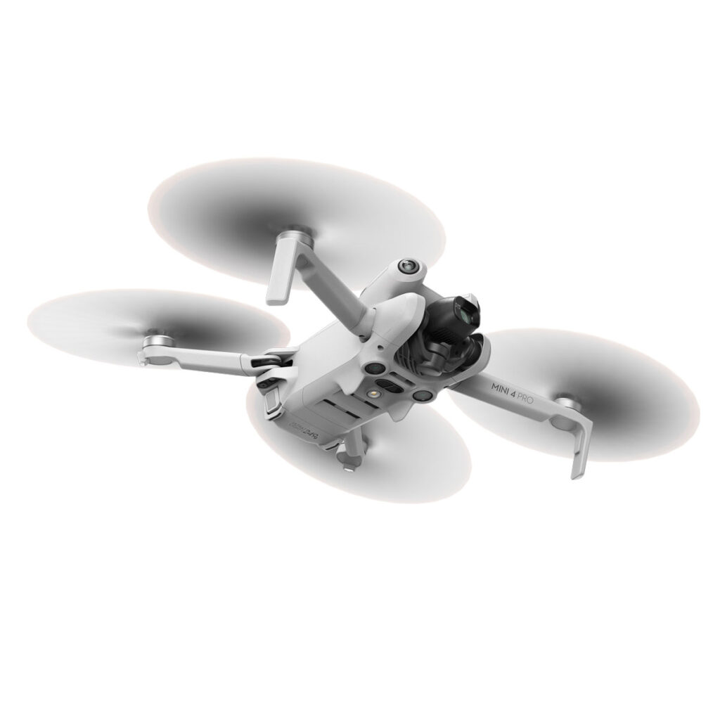 DJI_mini4pro_008 DJI Mini 4 Pro -lennokki, valkoinen nelikopteri, jossa on pyörivät potkurit, näkyy kesken lennon valkoista taustaa vasten. Etupuolen alapuolella on sen kamera ja anturit.