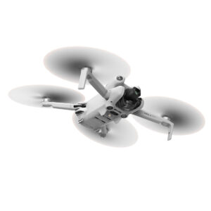 DJI_mini4pro_008 DJI Mini 4 Pro -lennokki, valkoinen nelikopteri, jossa on pyörivät potkurit, näkyy kesken lennon valkoista taustaa vasten. Etupuolen alapuolella on sen kamera ja anturit.