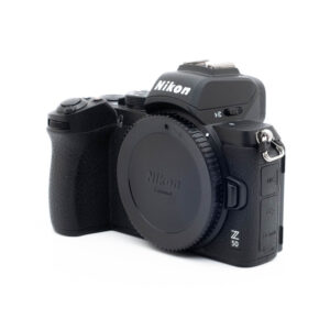 Nikon Z50-5-3 Nikon Z50 (SC 4000, Kunto K5) - Käytetty peilittömän kameran runko on oikealla, objektiivin suojus päällä, ja siinä näkyy kahva ja sivuportit.