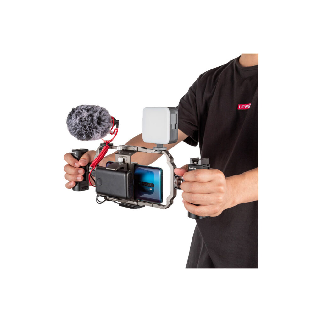 Smallrig_3384_005jpg Henkilö pitää kädessään SmallRig 3384 Professional Vlogging Kitiä, jossa on kaksi kahvaa, asennettu älypuhelin, mikrofoni, jossa on karvainen tuulisuoja, ja LED-valo - ihanteellinen videokuvaukseen tai vloggaamiseen.