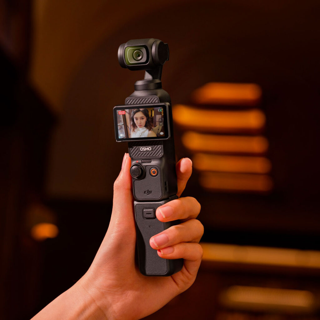 DJI_Osmo_3_002 In her hand is a DJI Osmo Pocket 3 battery grip attached to a camera, whose rotating screen shows a woman's face against a warmly lit, blurred background in shades of orange and brown.