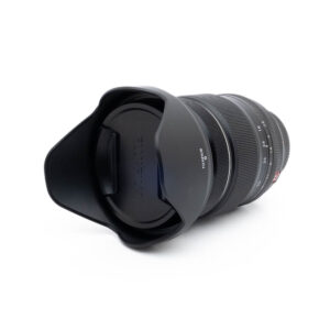 Fujinon 16-55mm f28 R LM WR-3 Fujinon GF 50mm f/3.5 R LM WR (sis.ALV24%) - Käytetty musta kameraobjektiivi, johon on kiinnitetty vastavalosuoja ja suojus, näkyy valkoisella taustalla, ja siinä näkyy objektiivin etupuoli ja osa objektiivin piipusta.