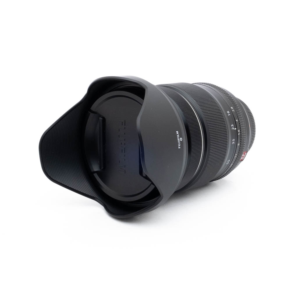 Fujinon 16-55mm f28 R LM WR-3 Fujinon GF 50mm f/3.5 R LM WR (sis.ALV24%) - Käytetty musta kameraobjektiivi, johon on kiinnitetty vastavalosuoja ja suojus, näkyy valkoisella taustalla, ja siinä näkyy objektiivin etupuoli ja osa objektiivin piipusta.