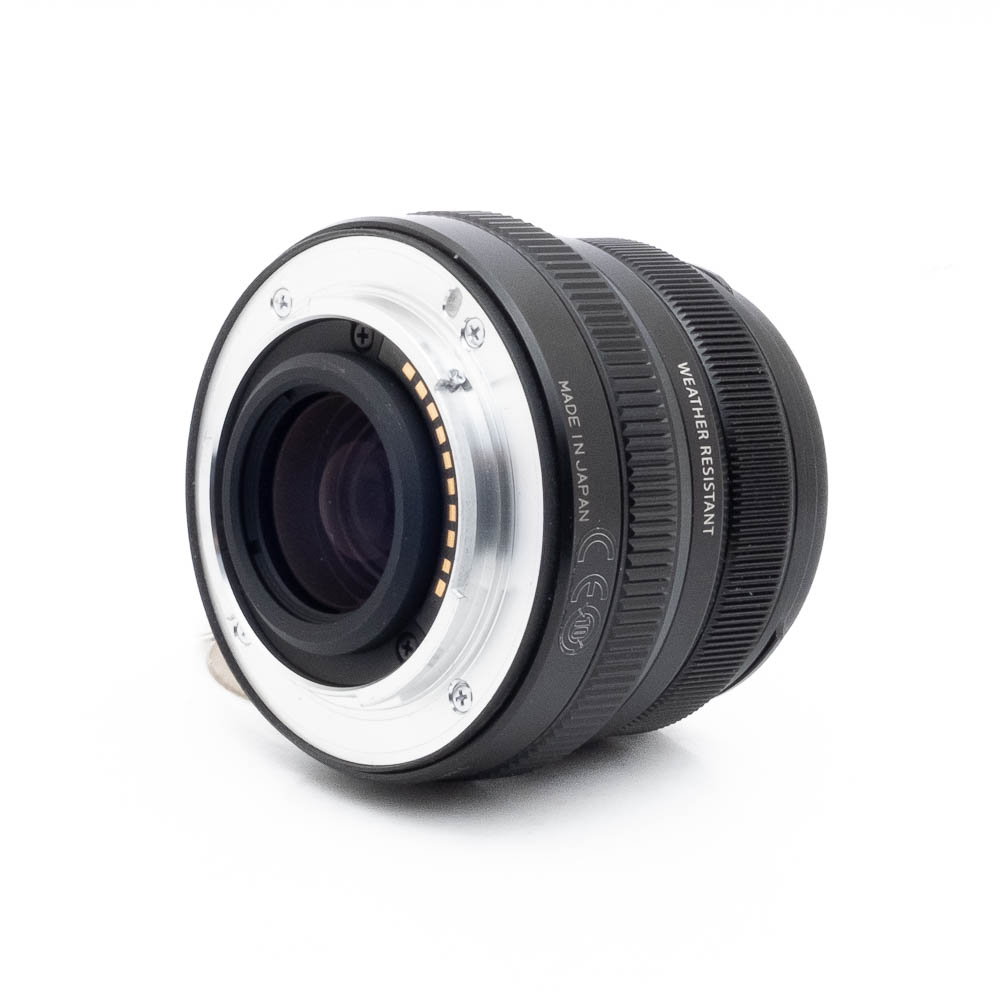 Fujinon 35mm f2 R WR-1 Fujinon GF 50mm f/3.5 R LM WR (sis.ALV24%) - Käytetty objektiivi, musta, hopeisella metallikiinnityksellä, lepää valkoisella pinnalla, joka on käännetty taaksepäin, jotta elektroniset kontaktit ja säänkestävyysmerkintä tulevat esiin.