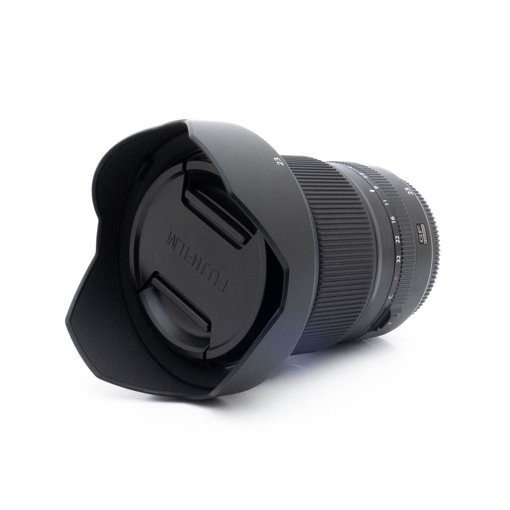 Fujinon GF 23mm f4 R LM WR-3 Fujinon GF 50mm f/3.5 R LM WR (sis.ALV24%) - Käytetty kameran objektiivi, johon on kiinnitetty vastavalosuoja ja suojus, makaa kyljellään valkoista taustaa vasten.