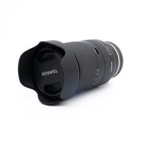 Tamron 28-200mm f/2.8-5.6 Di III RXD Sony E - Käytetty objektiivi, jossa on vastavalosuoja ja suojus, makaa kyljellään valkoista taustaa vasten.