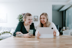 Kaksi naista istuu puupöydän ääressä hymyillen ja katselee tablettia ja älypuhelinta valoisassa ja modernissa sisätilassa.