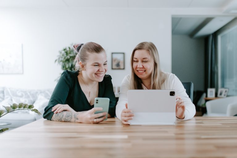 Kaksi naista istuu puupöydän ääressä hymyillen ja katselee tablettia ja älypuhelinta valoisassa ja modernissa sisätilassa.
