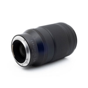Nikon Z MC 105mm f28 S-1 Nikon Nikkor Z MC 105mm f/2.8 VR S (Kunto K5) - Käytetty musta kameraobjektiivi makaa kyljellään, ja siinä näkyy metallinen objektiivin kiinnitys ja elektroniset kontaktit toisessa päässä valkoista taustaa vasten.