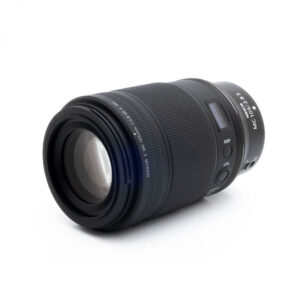 Nikon Z MC 105mm f28 S-2 Musta Nikon Nikkor Z MC 105mm f/2.8 VR S (Kunto K5) - Käytetty makro-objektiivi makaa kyljellään valkoista taustaa vasten, ja siinä on kuvioidut kahvarenkaat ja valkoiset kirjaimet.