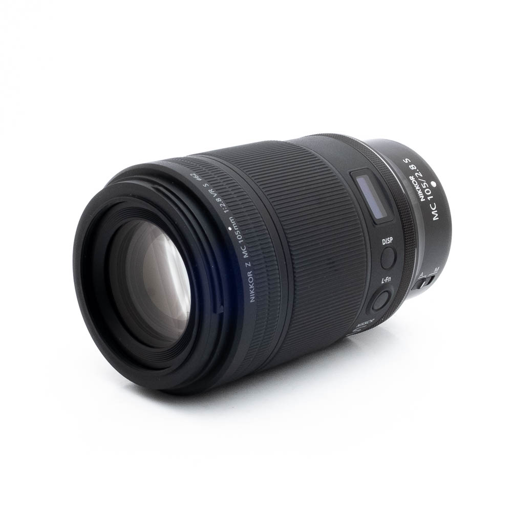 Nikon Z MC 105mm f28 S-2 Musta Nikon Nikkor Z MC 105mm f/2.8 VR S (Kunto K5) - Käytetty makro-objektiivi makaa kyljellään valkoista taustaa vasten, ja siinä on kuvioidut kahvarenkaat ja valkoiset kirjaimet.