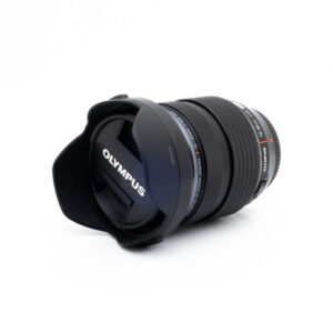 Olympus 12-40mm f28 PRO-3-2 Nikon Nikkor Z MC 105mm f/2.8 VR S (Kunto K5) -objektiivi, johon on kiinnitetty vastavalosuoja, makaa kyljellään valkoista taustaa vasten. Objektiivin suojuksessa on selvästi näkyvissä tuotemerkki NIKON.