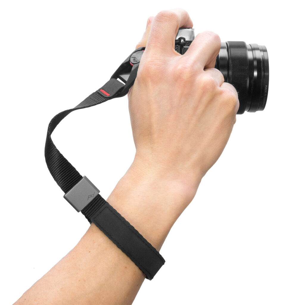 Peak-Design-Cuff-Black-2 Käsi pitää kameraa Peak Design Cuff Rannehihna - musta rannehihna, joka on kiinnitetty turvallisesti ranteen ympärille valkoisella pohjalla.